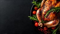 Roasted whole chicken with herbs, spices and cherry tomatoes on black plate against dark background, festive dinner composition with copy space.