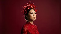 Asian woman wearing elaborate red beaded garment and ornate golden spiky crown headdress against deep burgundy background. Fashion and editorial use.