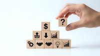 Hand placing wooden cube with question mark over business icons pyramid representing decision making and financial planning strategy.