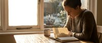 Young woman reading book by window with warm sunlight streaming in, cozy morning study session at home with coffee cup on wooden table for relaxation content.