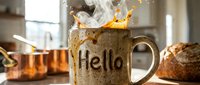 Steaming ceramic mug with Hello inscription and dynamic coffee splash on kitchen counter with bread and copper pots in background.