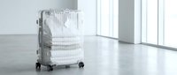 Transparent hard-shell carry-on suitcase packed with white folded clothes standing on concrete floor in bright minimalist interior with large windows.