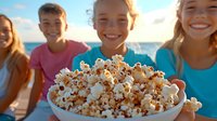 Smiling diverse group of young friends sharing fresh popcorn outdoors at beach on sunny summer day, focus on snack bowl in foreground against ocean backdrop.