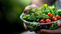 Fresh green salad with cherry tomatoes and basil leaves in glass bowl held by female hands against blurred natural background, healthy eating concept.
