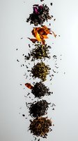 Variety of loose tea leaves arranged vertically on white background, showing different types and colors from black to green, with scattered dried petals and herbs.