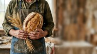 Baker or artisan holding freshly baked sourdough bread loaf with wheat stalks in rustic workshop setting, showcasing traditional breadmaking craft and quality ingredients.