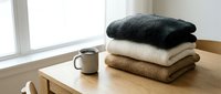 Stacked cozy knit sweaters in charcoal, cream and tan on wooden table with mug by window for home decor and lifestyle content.