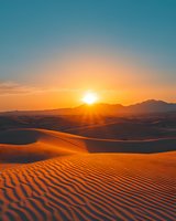 Golden sunset over rippled sand dunes in desert landscape, dramatic mountain silhouettes against orange sky and sun rays creating deep shadows on sandy terrain.