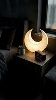 Modern crescent moon table lamp glowing warmly on wooden nightstand beside bed in contemporary bedroom interior with ambient lighting and cozy atmosphere.