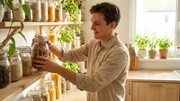 Man in casual beige shirt examining glass jar of dried pasta in bright kitchen with shelves of pantry staples and potted herbs, sustainable food storage and organization.