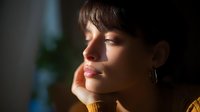 Young woman in yellow sweater with thoughtful expression illuminated by soft natural window light, resting head on hand in contemplative pose against dark background.