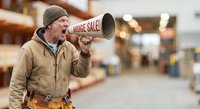 Mature Caucasian male warehouse worker shouting through megaphone announcing huge sale in industrial storage facility with shelving background.