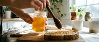 Unrecognizable person spreading honey on whole grain bread with a wooden brush in bright kitchen with potted plants and natural light.