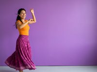 Young woman with dark hair and medium skin tone wearing yellow crop top and purple patterned skirt, flexing arm muscle in confident pose against purple background for fitness, wellness, empowerment.