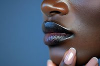 Close-up profile of dark-skinned woman wearing metallic blue and silver lipstick, finger with pink manicure touching chin against blue background. Fashion beauty concept.