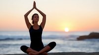 Woman practicing yoga in lotus position with prayer hands at sunset on beach, silhouetted against golden sky and ocean waves.