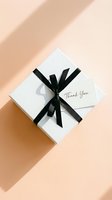 Elegant white gift box tied with black satin ribbon and thank you card on pastel peach background, top view for holiday or celebration.