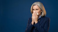 Mature woman with gray hair wearing dark blazer holding tissue to face against blue background, depicting cold, flu, or allergy symptoms for healthcare and wellness content.