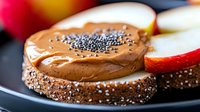 Creamy peanut butter spread on multigrain bread topped with chia seeds and fresh apple slices, close-up view on dark background. Healthy breakfast concept.