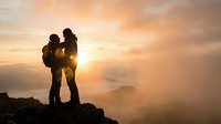 Silhouette of two hikers with backpacks standing on mountain peak at golden sunrise, embracing scenic valley view for adventure and travel concepts.