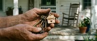 Weathered hands holding house keys with a wooden home keychain on a porch background, symbolizing real estate ownership and property purchase.