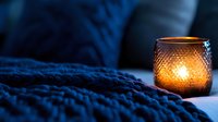 Glowing candle in textured glass holder against cozy blue knitted blanket creating warm atmospheric light for relaxation and mindfulness practices.