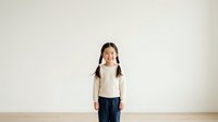 Young Asian girl with braided hair wearing cream sweater and navy skirt stands against white wall, suitable for education, childhood, and lifestyle marketing materials.