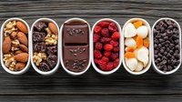 Assorted healthy snacks in white ceramic containers including nuts, dark chocolate, dried fruits, and chocolate chips on rustic wooden background.