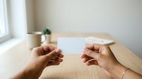 Hands of two people exchanging blank white business card at wooden table with plant and cup, suitable for corporate branding and networking concepts.