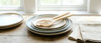 Stacked ceramic plates with wooden spoon and fork on rustic wooden table near window, natural linen napkin beside, soft light.