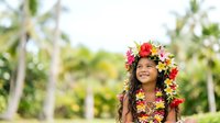 Young girl with long dark hair and medium skin tone wearing a vibrant floral lei and matching Hawaiian shirt, smiling outdoors in tropical garden setting with palm trees and lush greenery.