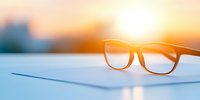 Modern eyeglasses on light surface against blurred sunset background with golden sun flare, minimalist composition for optical business and vision care.