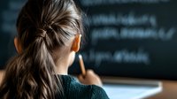 Young female student with brown hair in ponytail looking at chalkboard with mathematical formulas, rear view in classroom during studies. Focus on hair and blurred background.