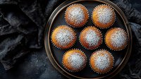 Fresh homemade vanilla cupcakes dusted with powdered sugar arranged on dark rustic plate against black textile background, overhead view of baked treats.