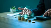 Professional businessman analyzing real estate investment data with digital house hologram and coin stacks on modern desk for financial planning.