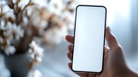 Hand holding modern smartphone with blank white screen against blurred background with cotton flowers in vase, mockup for app or website design presentation.