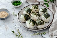 Rice balls wrapped in nori seaweed, topped with sesame seeds and fresh herbs on ceramic plate. Traditional Asian cuisine served with green onions and sesame.