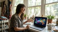 Young woman with dark hair and light skin tone, wearing a patterned shirt, sitting at a desk by bright windows, working on laptop displaying colorful digital graphics, surrounded by plants and creative supplies.