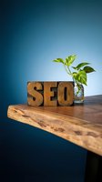 Wooden letterpress blocks spelling SEO on a rustic table beside a green plant in a glass vase, blue background for digital marketing concepts.