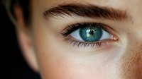 Close-up macro shot of bright blue human eye with freckles and dark eyebrow. Natural sunlight highlights iris detail and skin texture. Artistic portrait photography.