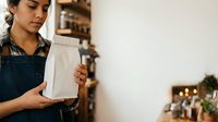 Female barista with South Asian ethnicity holding white paper bag in coffee shop, ready for customer order or takeout service.