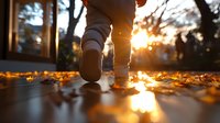 Person walking on fallen autumn leaves during golden sunset hour, low angle view showing shoes and legs with warm sunlight creating magical backlit glow.