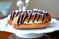 Fresh baked chocolate eclair with vanilla cream filling and dark chocolate drizzle on white serving plate, close up view of French pastry dessert.