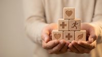Hands holding wooden blocks with healthcare symbols including hearts, medical cross, and wellness icons representing holistic health concept.