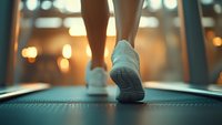 Close-up view of woman's feet in white sneakers running on treadmill at gym with warm bokeh lights in background, focus on shoe sole during workout.