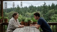 Young couple enjoying morning coffee together on rustic wooden deck surrounded by lush green forest and potted herbs creating peaceful outdoor lifestyle moment.