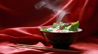 Steaming hot Asian soup in black bowl with fresh herbs and chopsticks on red silk background, dramatic food photography with rising steam effect.
