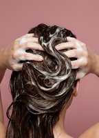 Woman washing long brown hair with shampoo foam creating bubbles during hair care routine against pink background for beauty and hygiene concepts.