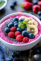 Pink smoothie bowl topped with fresh blueberries, raspberries, banana slices, chia seeds and mint leaf garnish on dark wooden surface, healthy breakfast concept.