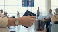 Young adult with medium skin tone holding passport at airport terminal with travelers in background, travel documentation and immigration concept.